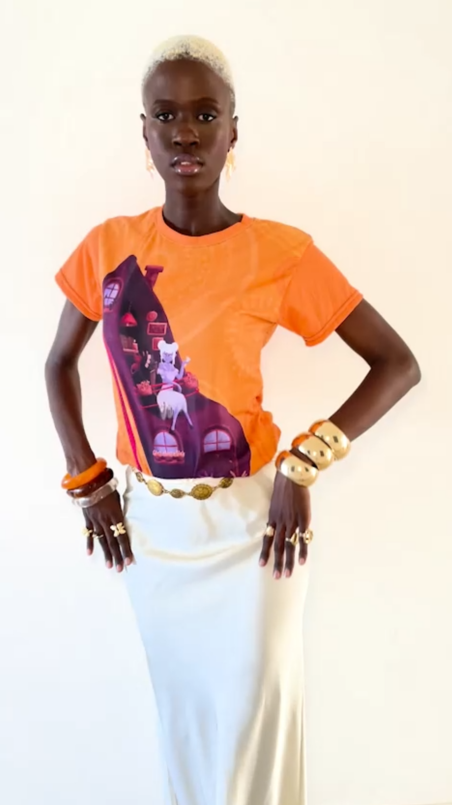 Person wearing an orange shirt with a graphic design and white skirt on a plain background
